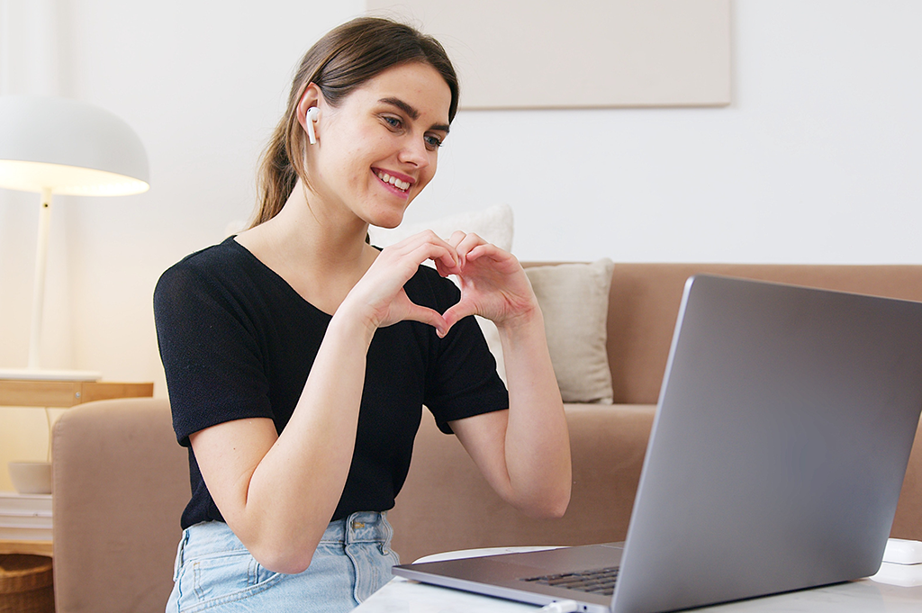 A woman makes a heart sign with her hands to her computer camera.